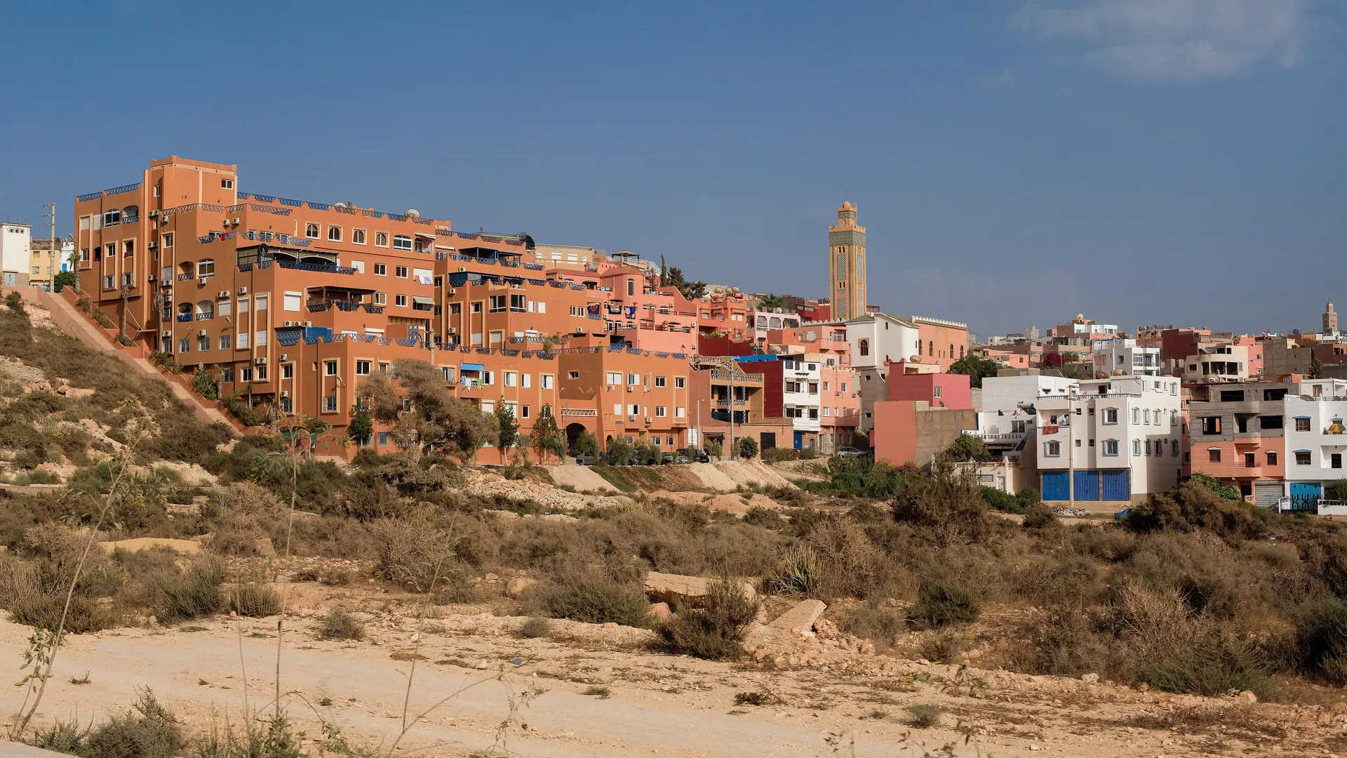 Panorama du village de Tamraght depuis l'oued — maisons colorées, minaret, et la plage de Banana au-delà