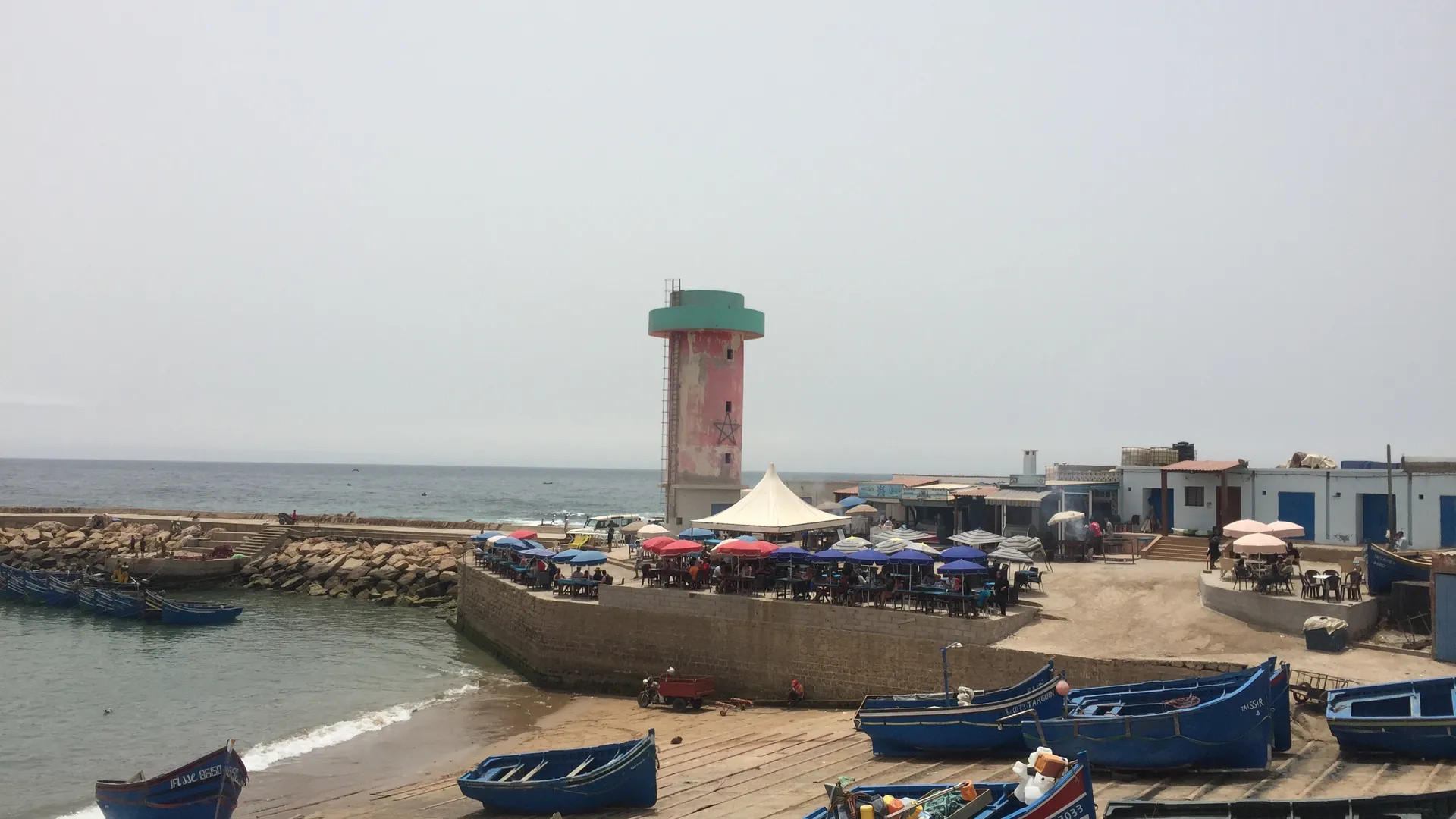 Barques de pêche bleues alignées dans le port d'Imsouane — le phare et les cafés en arrière-plan